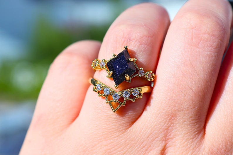 May include: Two gold rings are displayed on a finger. The top ring features a dark blue, square-cut gemstone. The second ring is a chevron shape with small, clear gemstones. Both rings have intricate gold detailing.