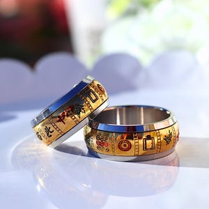 May include: Two stainless steel rings with gold-colored bands. The bands feature red, green, and black decorative elements. The rings are on a white surface, with a blurred background of white and green.