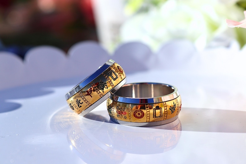 May include: Two stainless steel rings with gold-colored bands. The bands feature red, green, and black decorative elements. The rings are on a white surface, with a blurred background of white and green.