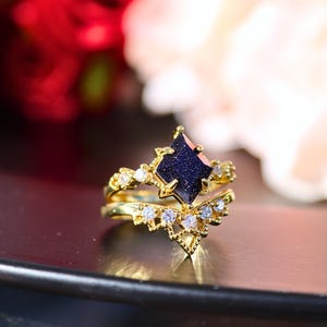 May include: A gold-colored ring set featuring a dark blue gemstone in a diamond shape, surrounded by smaller white gemstones. The set includes a matching band with a V-shaped design, also adorned with white gemstones.
