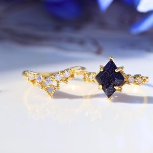 May include: Two gold rings. One ring features a dark blue, square-cut stone, and the other has a curved design with small, clear stones. Both rings have a delicate, ornate design, and are set against a white background.