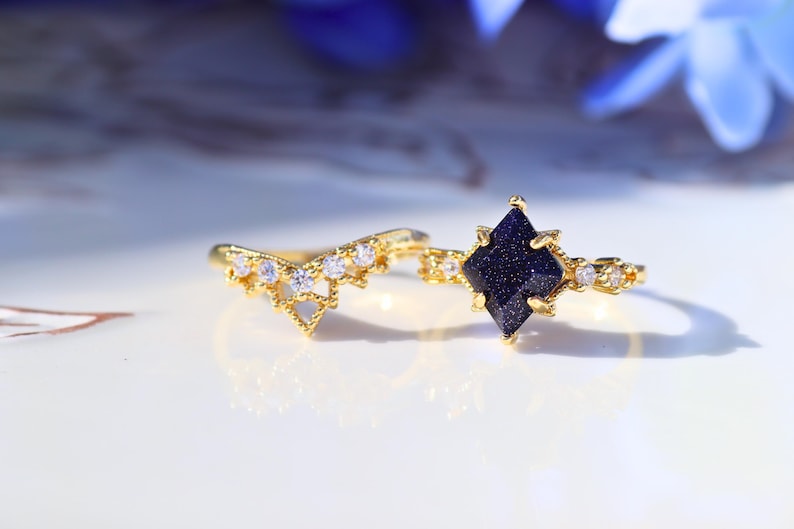 May include: Two gold rings. One ring features a dark blue, square-cut stone, and the other has a curved design with small, clear stones. Both rings have a delicate, ornate design, and are set against a white background.