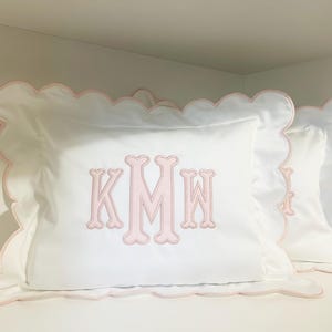 May include: A white rectangular pillowcase featuring a scalloped edge trimmed in light pink. The center of the pillowcase displays a large, embroidered monogram with the letters KMW in a light pink thread with a decorative, rounded font style.