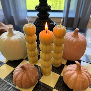 May include: A still-life arrangement featuring pumpkin-shaped candles and decorative pumpkins. Three stacked pumpkin candles are lit, casting a warm glow. The pumpkins are in shades of orange, peach, and white, set on a black and white checkered surface.