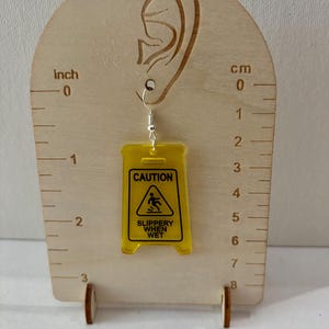 Puede incluir: Un solo pendiente amarillo de acr&iacute;lico con forma de se&ntilde;al de "Precauci&oacute;n: Suelo resbaladizo". El pendiente cuelga de un gancho plateado. El pendiente mide aproximadamente 5 cm de largo.