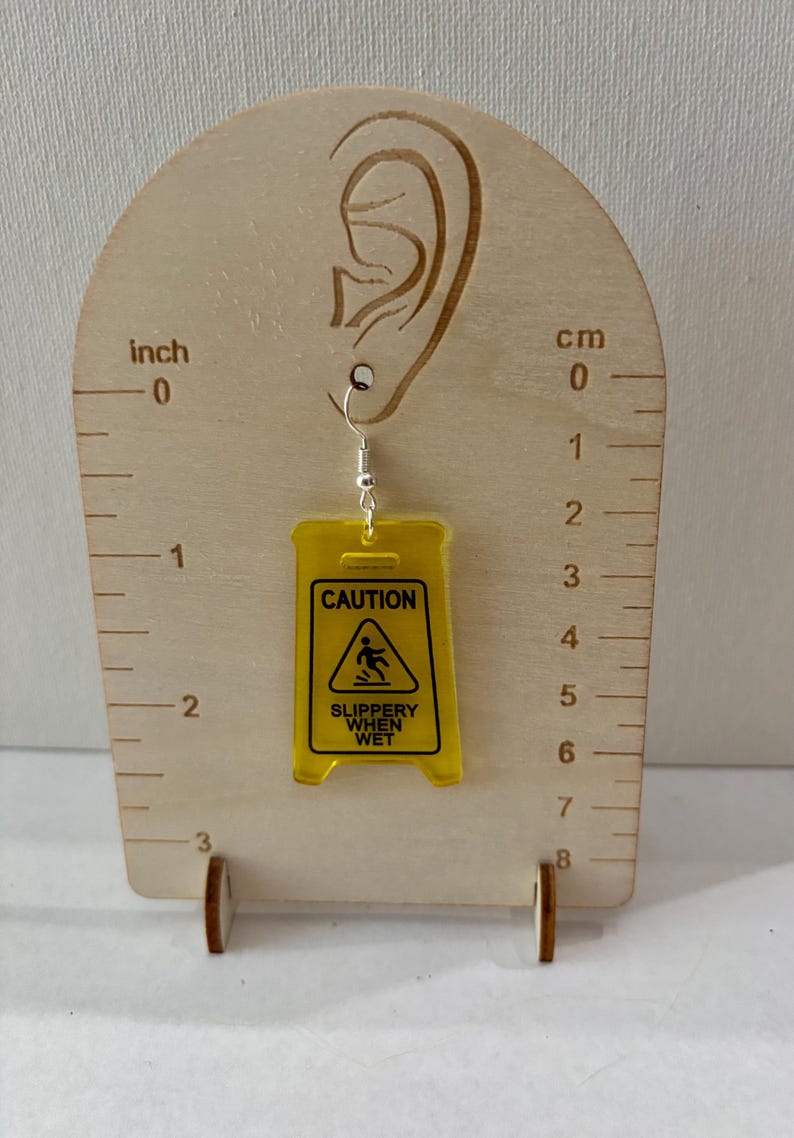 Puede incluir: Un solo pendiente amarillo de acr&iacute;lico con forma de se&ntilde;al de "Precauci&oacute;n: Suelo resbaladizo". El pendiente cuelga de un gancho plateado. El pendiente mide aproximadamente 5 cm de largo.