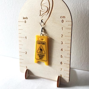 Puede incluir: Un pendiente amarillo con forma de se&ntilde;al de "Precauci&oacute;n: Suelo resbaladizo". El pendiente est&aacute; suspendido de un gancho para la oreja. El pendiente mide aproximadamente 2,5 cm de largo.