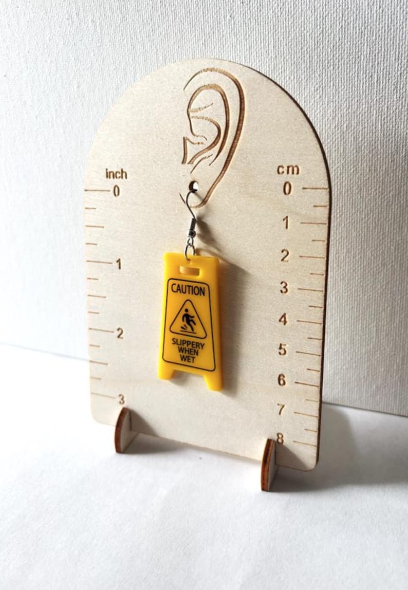 Puede incluir: Un pendiente amarillo con forma de se&ntilde;al de "Precauci&oacute;n: Suelo resbaladizo". El pendiente est&aacute; suspendido de un gancho para la oreja. El pendiente mide aproximadamente 2,5 cm de largo.