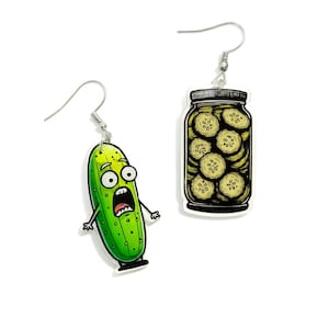 May include: A pair of novelty earrings featuring a green pickle character with a shocked expression and a jar of pickle slices. The earrings have silver-colored fish hook closures and are set against a white background.