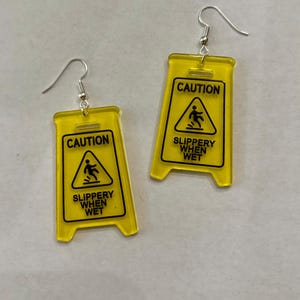 Puede incluir: Un par de pendientes novedosos con forma de se&ntilde;ales de precauci&oacute;n amarillas. Cada pendiente presenta las palabras "CAUTION" encima de un gr&aacute;fico de una persona resbalando, con "SLIPPERY WHEN WET" debajo. Los pendientes tienen cierres de gancho de pez plateados.