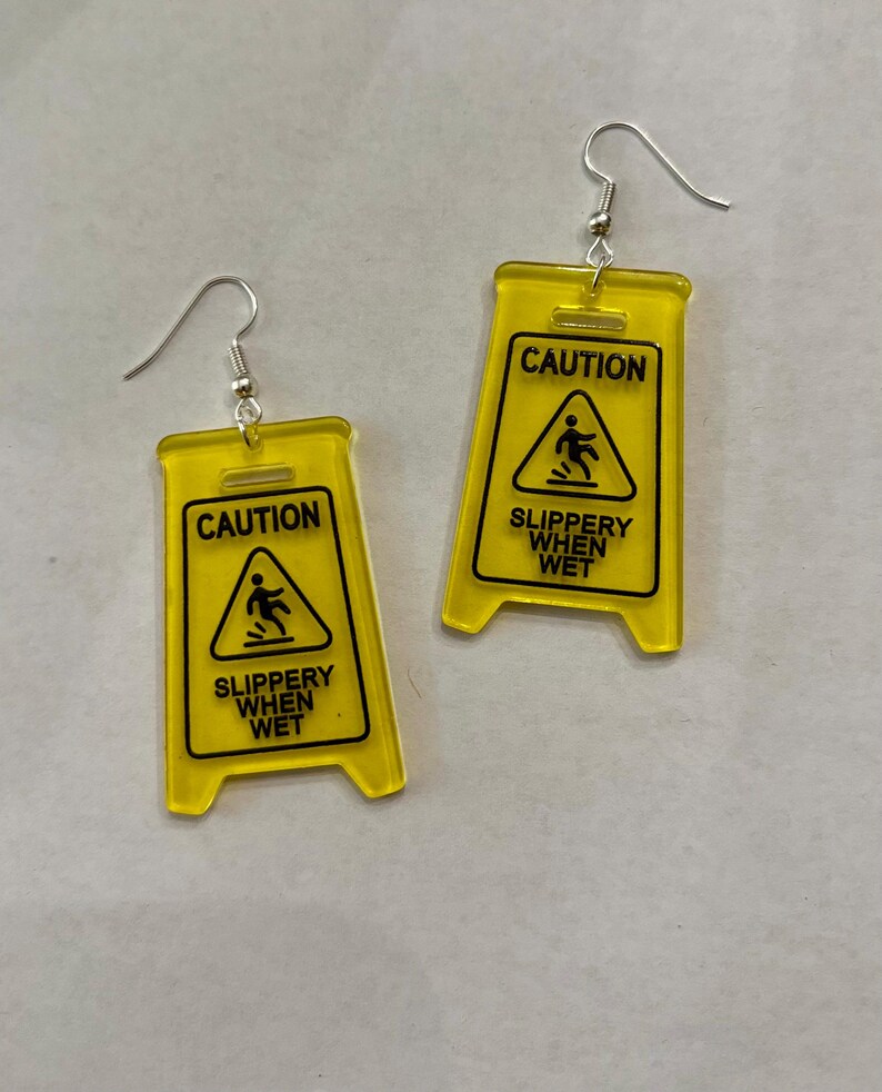 Puede incluir: Un par de pendientes novedosos con forma de se&ntilde;ales de precauci&oacute;n amarillas. Cada pendiente presenta las palabras "CAUTION" encima de un gr&aacute;fico de una persona resbalando, con "SLIPPERY WHEN WET" debajo. Los pendientes tienen cierres de gancho de pez plateados.