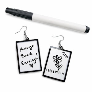 Może przedstawiać: Kolczyki w stylu tablicy z czarnymi ramkami, jeden z napisem "Message Board Earrings!" i sercem, drugi z rysunkiem kwiatu i słońca. Biało-czarny marker znajduje się nad nimi.