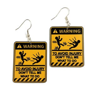 Puede incluir: Un par de pendientes novedosos con un diseño de señal de advertencia amarilla y negra. Los pendientes presentan el texto "WARNING" y "TO AVOID INJURY DON'T TELL ME WHAT TO DO". Los pendientes tienen cierres de gancho de pez plateados.