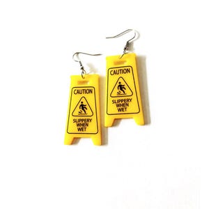 Puede incluir: Un par de pendientes novedosos con forma de se&ntilde;ales de precauci&oacute;n amarillas. Cada pendiente presenta las palabras "CAUTION" y "SLIPPERY WHEN WET" en texto negro, junto con un gr&aacute;fico de una persona resbalando. Los pendientes tienen cierres de gancho plateados.