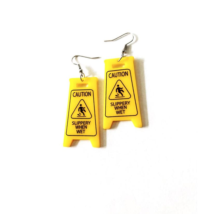 Puede incluir: Un par de pendientes novedosos con forma de se&ntilde;ales de precauci&oacute;n amarillas. Cada pendiente presenta las palabras "CAUTION" y "SLIPPERY WHEN WET" en texto negro, junto con un gr&aacute;fico de una persona resbalando. Los pendientes tienen cierres de gancho plateados.