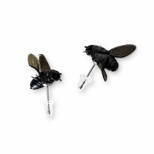 May include: A pair of stud earrings shaped like black bees with detailed wings and bodies. Each earring features a thin metal post and a clear plastic backing, designed to resemble the insect in flight with a dark, matte finish on the bee figures.