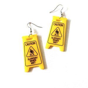 Puede incluir: Un par de pendientes novedosos con forma de se&ntilde;ales de precauci&oacute;n amarillas. Cada pendiente tiene las palabras "CAUTION" y "SLIPPERY WHEN WET" impresas en negro. Los pendientes tienen ganchos para la oreja de color plateado.