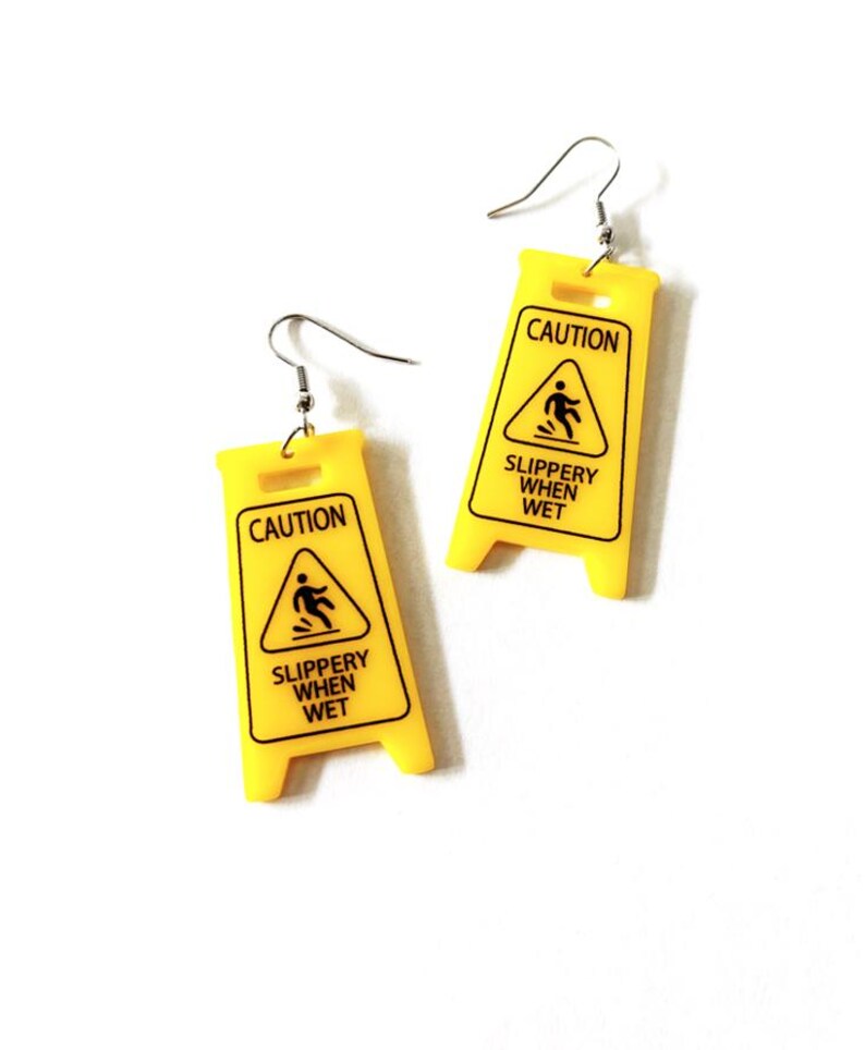 Puede incluir: Un par de pendientes novedosos con forma de se&ntilde;ales de precauci&oacute;n amarillas. Cada pendiente tiene las palabras "CAUTION" y "SLIPPERY WHEN WET" impresas en negro. Los pendientes tienen ganchos para la oreja de color plateado.