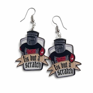 Peut inclure: Paire de boucles d'oreilles fantaisie avec un chevalier de dessin animé en armure noire et des accents rouges. Le chevalier est placé sur un fond blanc avec une bannière qui dit "tis but a scratch". Crochets argentés.