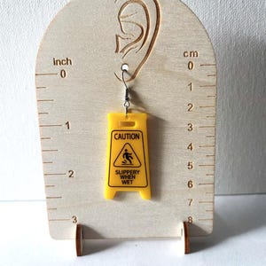 Puede incluir: Un pendiente de acr&iacute;lico amarillo con forma de se&ntilde;al de precauci&oacute;n, con el texto "CAUTION SLIPPERY WHEN WET". El pendiente est&aacute; suspendido de un gancho plateado. El pendiente mide aproximadamente 4,4 cm de largo.