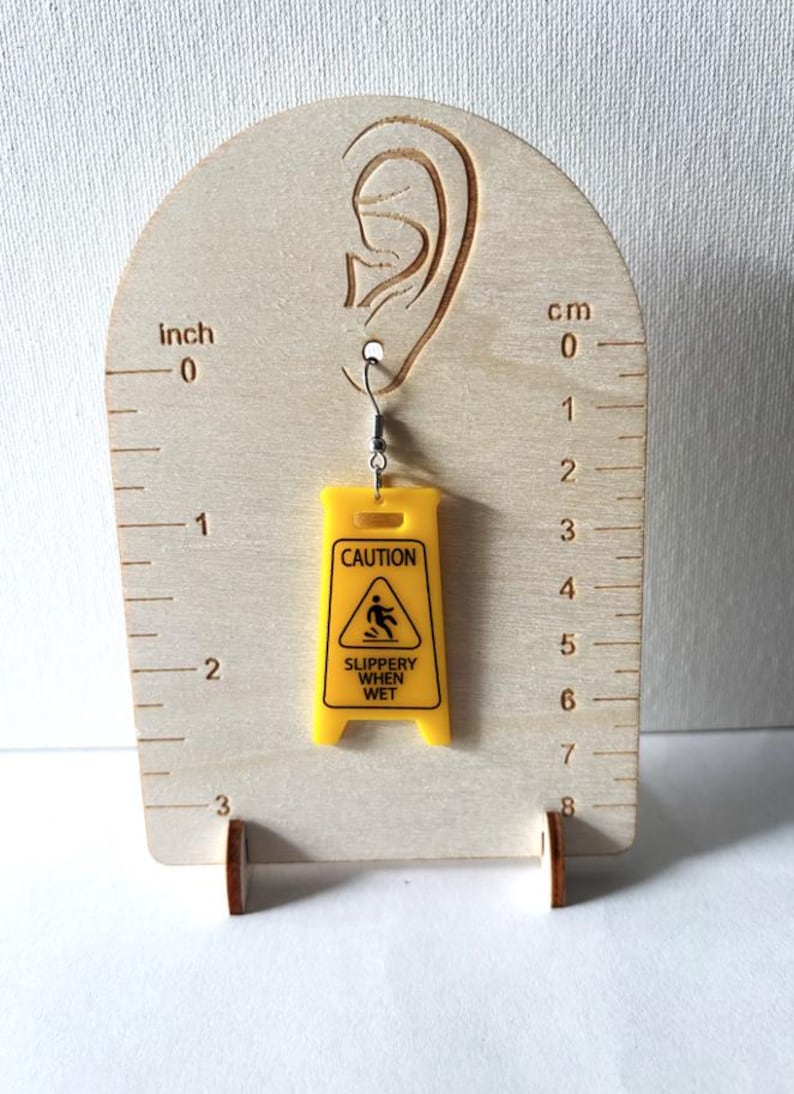 Puede incluir: Un pendiente de acr&iacute;lico amarillo con forma de se&ntilde;al de precauci&oacute;n, con el texto "CAUTION SLIPPERY WHEN WET". El pendiente est&aacute; suspendido de un gancho plateado. El pendiente mide aproximadamente 4,4 cm de largo.