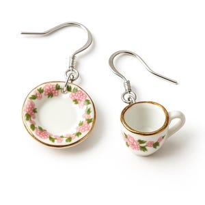 May include: A pair of dangle earrings featuring a miniature teacup and saucer design. The white porcelain pieces are adorned with pink floral patterns and gold trim. The earrings hang from silver-colored hooks.