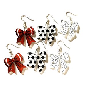 May include: Six pairs of bow-shaped earrings. Two pairs are red with white accents, two pairs have a soccer ball pattern, and two pairs are outlined in white. All earrings have silver-colored fish hook closures.