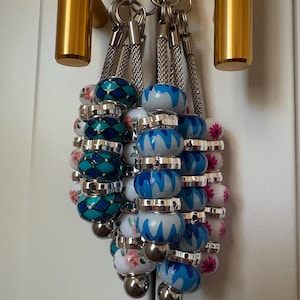 May include: A collection of beaded keychains with silver-tone metal accents. The beads feature various designs, including floral patterns, geometric shapes, and wavy blue patterns. Each keychain has a silver-tone metal clasp.