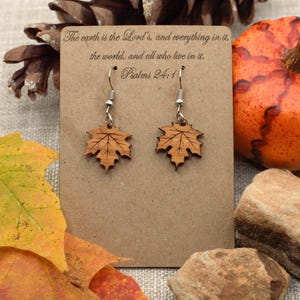 May include: A pair of wooden leaf earrings with silver hooks, displayed on a brown card with the text "The earth is the Lord's, and everything in it, the world, and all who live in it. Psalms 24:1". The earrings are set against a backdrop of fall foliage and a pumpkin.