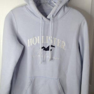 May include: A light blue hooded sweatshirt featuring the white text HOLLISTER, a dark blue bird logo, and the word California in white script. The garment includes a front kangaroo pocket, long sleeves, and matching light blue drawstrings.