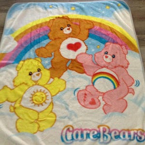 May include: A white Care Bears blanket featuring three cartoon bears in yellow, brown, and pink. The bears are set against a rainbow and cloud background, with the words "Care Bears" in blue.