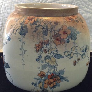 Puede incluir: Un jarrón de cerámica con cuerpo redondeado y boca ancha, que presenta una base de color crema decorada con un delicado patrón de flores naranjas, hojas azules y enredaderas colgantes. El borde superior tiene un suave degradado marrón.