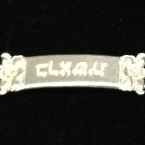 May include: A silver-toned metal bar with decorative floral-like embossed ends. The center of the bar features engraved text that appears to read CLXAM. The piece has a smooth, slightly curved surface and a metallic finish.