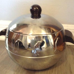 May include: A shiny, silver-colored metal ice bucket with a lid and a dark brown knob. The ice bucket has two handles and a decorative design on the side. The bucket is sitting on a wooden surface.