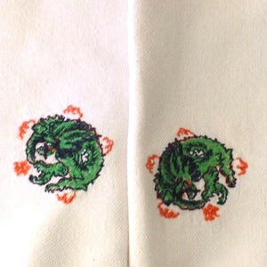 May include: Two off-white towels with embroidered green dragon designs. The dragons are surrounded by orange floral accents. The towels have orange trim along the bottom edge.