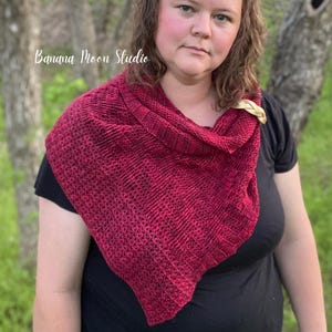 May include: A woman wearing a red and black knitted shawl with a black shirt. The shawl is draped around her neck and shoulders. The shawl has a textured pattern and is made of a soft yarn.