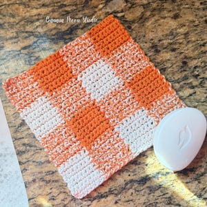 May include: A crocheted washcloth in orange and white, featuring a checkerboard design. A white bar of soap is placed beside it. The text "Banana Moon Studio" is visible in the upper left corner.