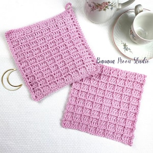 May include: Two pink crocheted pot holders with a square grid pattern. The pot holders are on a white surface. Banana Moon Studio is written in the background.