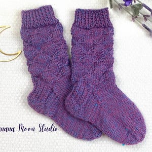 May include: A pair of hand-knitted socks in shades of purple and blue. The socks feature a textured pattern and ribbed cuffs. The text "Banana Moon Studio" is visible in the lower left corner.