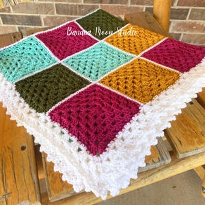 Pode incluir: Um cobertor de bebé em crochê com uma borda branca e quadrados em tons de turquesa, bordô, verde oliva e dourado. O cobertor é feito com um padrão de quadrado da avó.