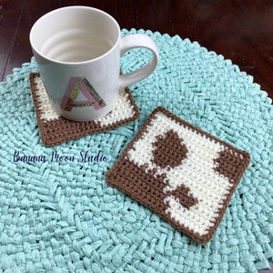 May include: Two crocheted coasters, one brown and white with a cow print design and the other brown and white with a letter A in the center. The coasters are on a light blue woven mat.
