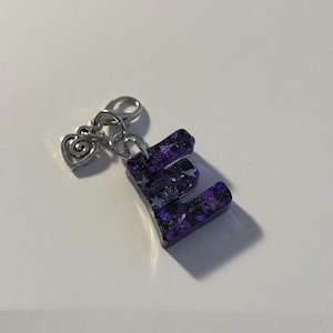 May include: A purple letter "E" keychain with silver hardware and a heart charm. The letter is filled with dark purple glitter and small star-shaped glitter pieces. The keychain is on a white surface.