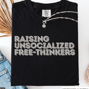 May include: Black t-shirt with the text "RAISING UNSOCIALIZED FREE-THINKERS" in a distressed, gray font. A silver necklace with a pendant is visible. The shirt is folded, with a tag from Comfort Colors visible at the neckline.