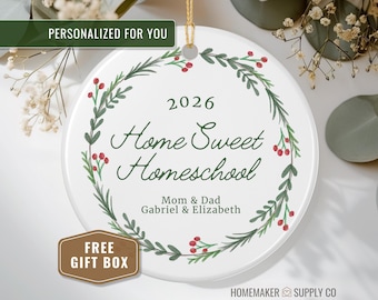 May include: A round white ceramic ornament featuring a green botanical wreath with red berries. The text reads 2026, Home Sweet Homeschool, Mom & Dad, and Gabriel & Elizabeth. A gold string is attached to the top for hanging the decorative piece.
