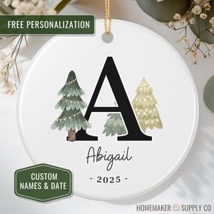 May include: White ceramic Christmas ornament with a gold string. The ornament features a black letter "A", watercolor pine trees, the name "Abigail", and the year "2025". The ornament has the text "FREE PERSONALIZATION" and "CUSTOM NAMES & DATE".