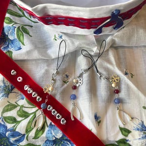 Puede incluir: Una cinta de terciopelo rojo con el texto "I love this song" en cuentas blancas, junto con tres colgantes decorativos con cuentas, herraduras y formas de trébol. El fondo presenta un gorro de marinero blanco y tela estampada floral.