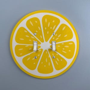 May include: A decorative light switch cover featuring a yellow lemon slice design with white accents. The cover has two white light switches and is mounted on a gray wall. The lemon design adds a bright, cheerful touch to any room.