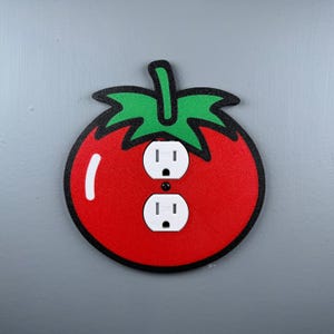 May include: A decorative red tomato-shaped light switch cover with a green stem and leaf design. The cover has two white electrical outlets and a black outline. The design is a fun and whimsical addition to any room.