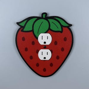 May include: A decorative strawberry-shaped wall plate with two white electrical outlets. The strawberry is red with black seeds and a green leafy top. The outline is black.