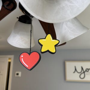 Peut inclure: Un ventilateur de plafond avec deux chaînes décoratives. L'une a un charme en forme de cœur rouge avec un reflet blanc, et l'autre a un charme en forme d'étoile jaune. Les pales du ventilateur sont marron.